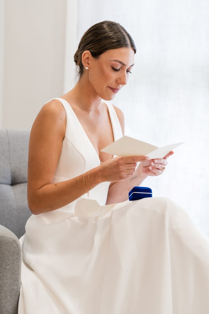 A bride in a white dress sits on a gray chair, reading a card with a blue ring box nearby. With her hair pulled back, she appears calm and focused in this elegant moment of editorial photography.