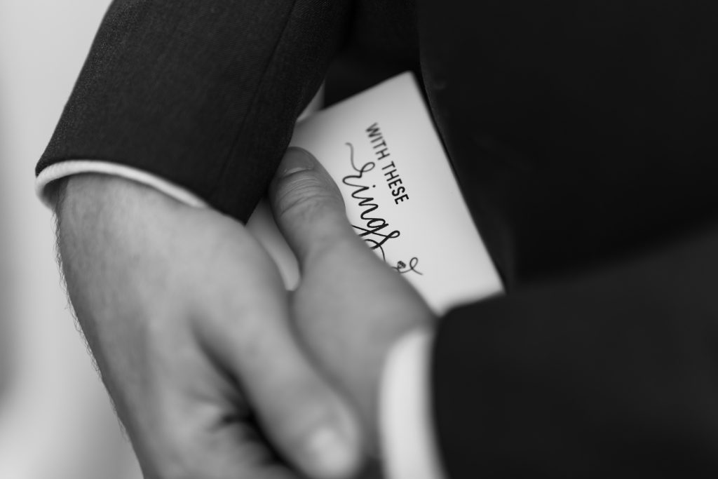 Close-up, black and white editorial photography of a person in a suit holding a card that reads WITH THESE rings, partially visible. The hand lightly grasps the card, suggesting a wedding or engagement setting in Toronto.