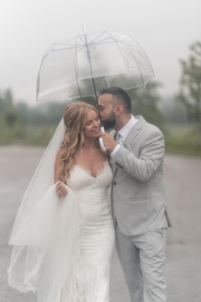 bride and groom go for a stroll on their wedding day in toronto as the groom kisses her on her cheek. it was a rainy day and an umbrella was a must!