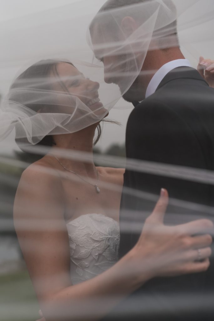 A bride and groom smile at each other closely under a sheer veil outdoors, their joy beautifully captured in a soft, romantic, documentary photography style—perfect for a dreamy Toronto wedding moment.