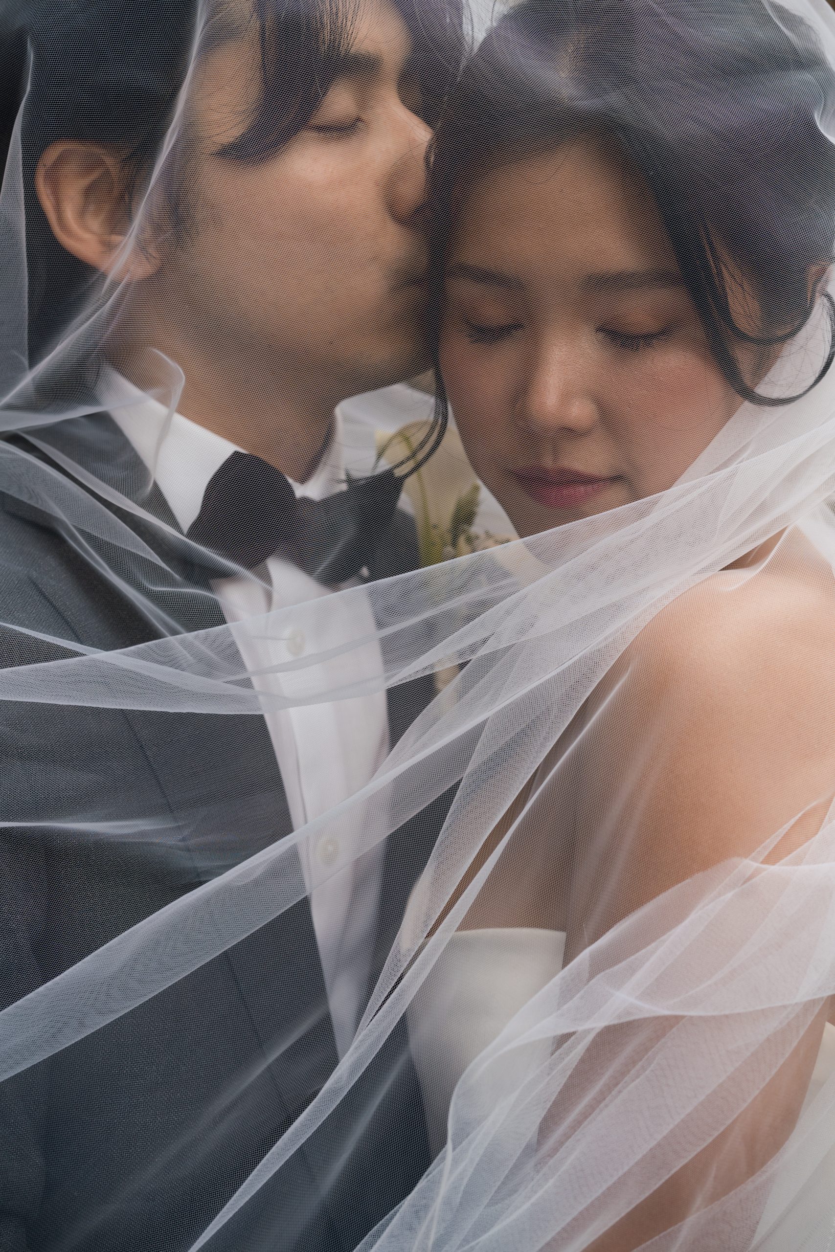 A groom in a suit gently kisses the forehead of his bride, both wrapped in a sheer white veil. Captured by a skilled photographer, this intimate and tender wedding moment feels timeless and editorial.