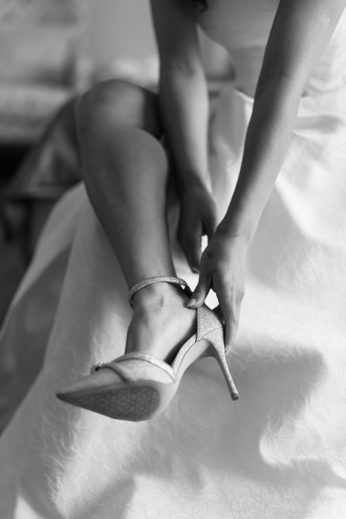 A woman in a white dress sits and fastens the ankle strap of a high-heeled shoe; the black and white, editorial-style image focuses on her hands, shoes, and legs.