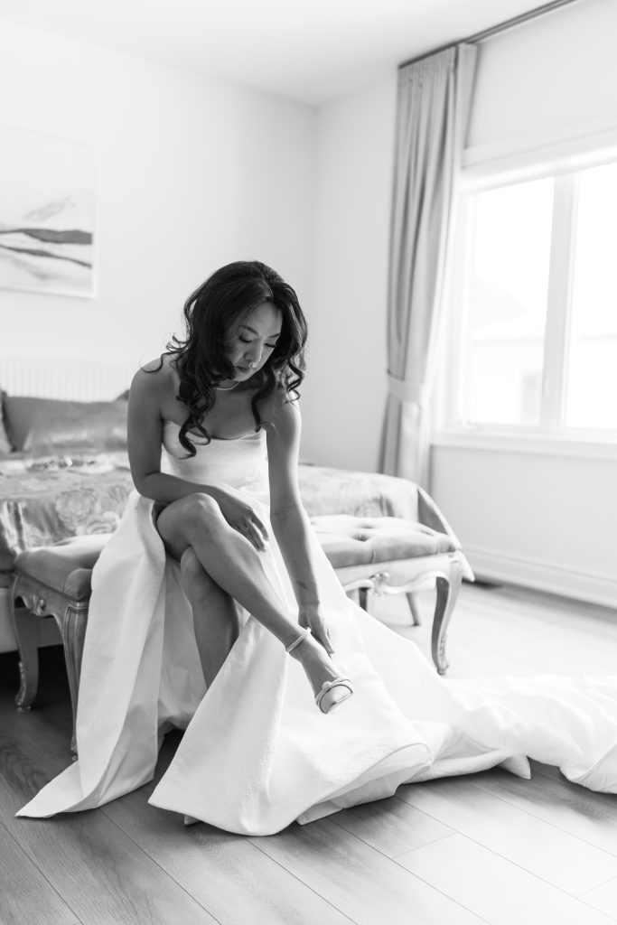 A bride in a white gown sits on a bench, adjusting her shoe in a bright, elegant room with large windows and curtains. Captured in black and white by a Toronto photographer with a documentary style.