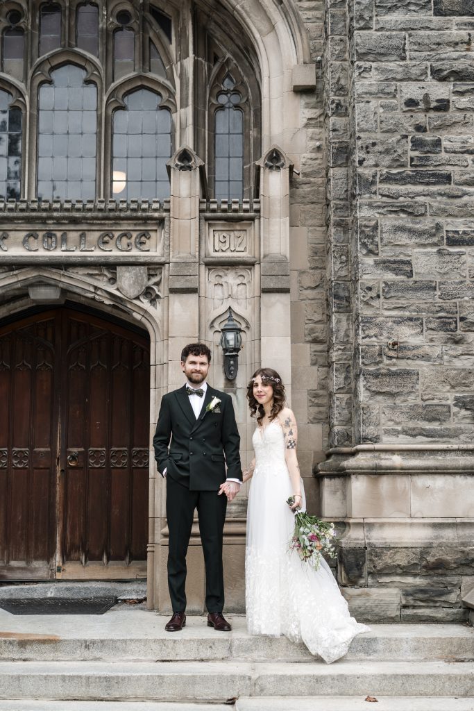 A bride in a white dress holding a bouquet and a groom in a dark suit stand hand in hand on stone steps outside a historic Toronto building with arched wooden doors and gothic windows, captured in an editorial style.