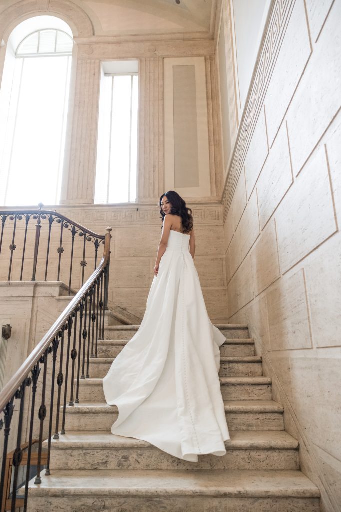 A woman in a long, elegant white dress stands on a marble staircase, looking back over her shoulder. Sunlight streams through tall windows, illuminating the grand interior in this stunning editorial photography scene.