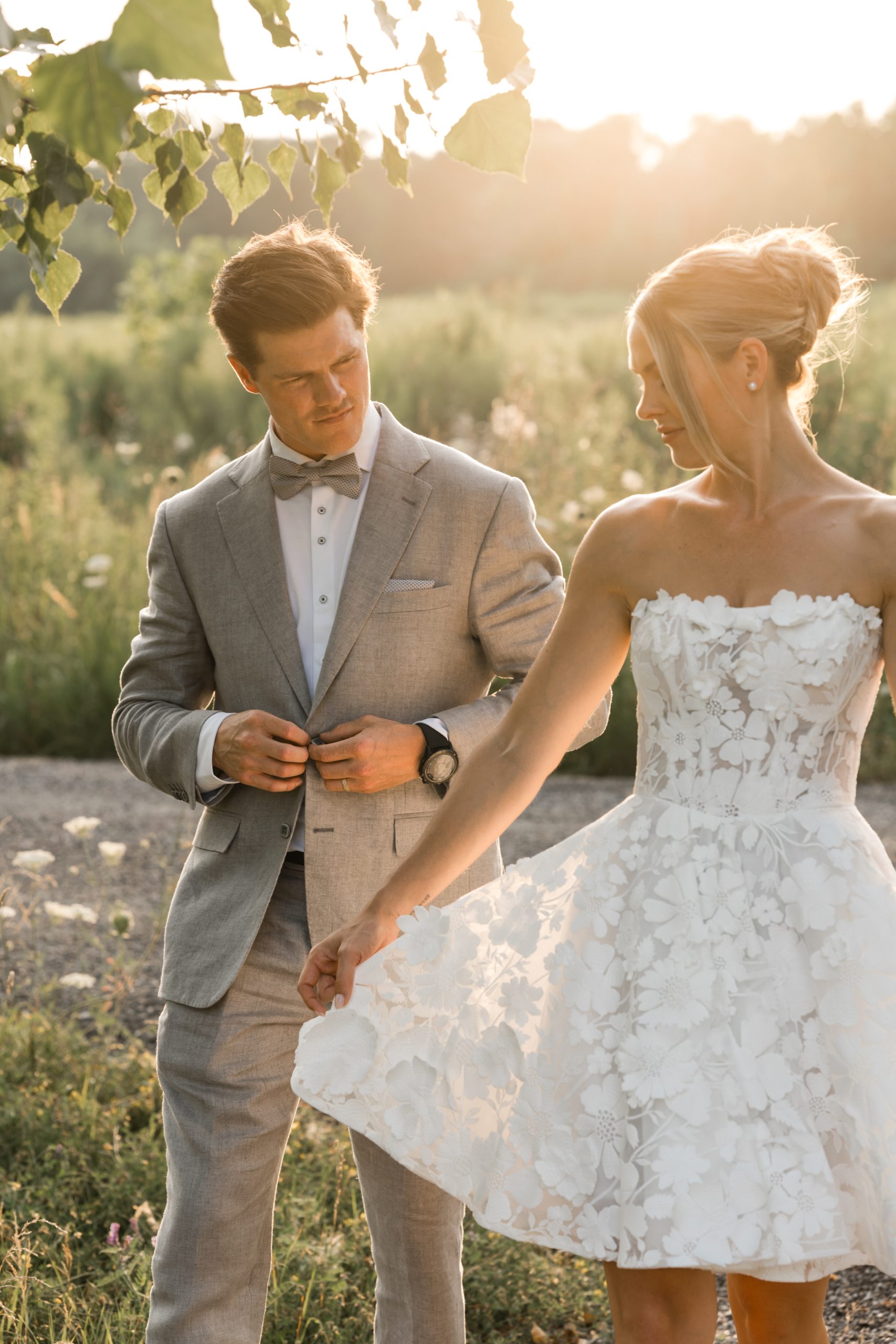 A bride in a strapless white floral dress and a groom in a light gray suit stand outdoors at sunset, surrounded by greenery—capturing the romance and charm of top Toronto Wedding Photography Locations.