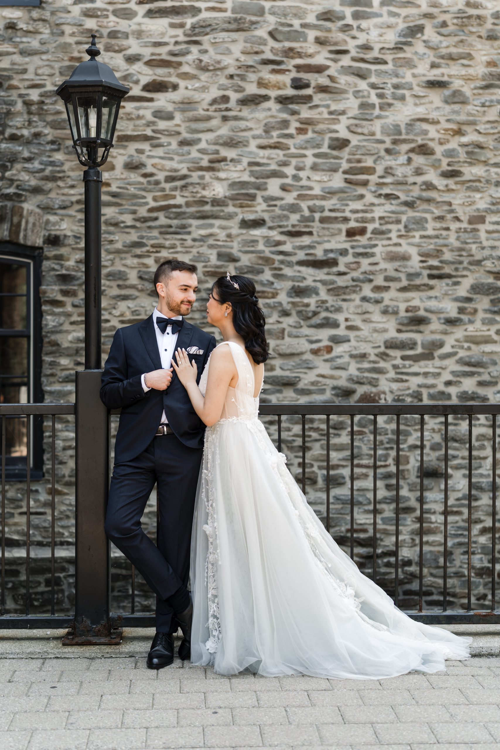 A bride in a white gown and a groom in a black tuxedo stand close together outdoors, smiling at each other by a wrought iron fence and old stone wall with a lamp post, captured in an editorial style by their wedding photographer.