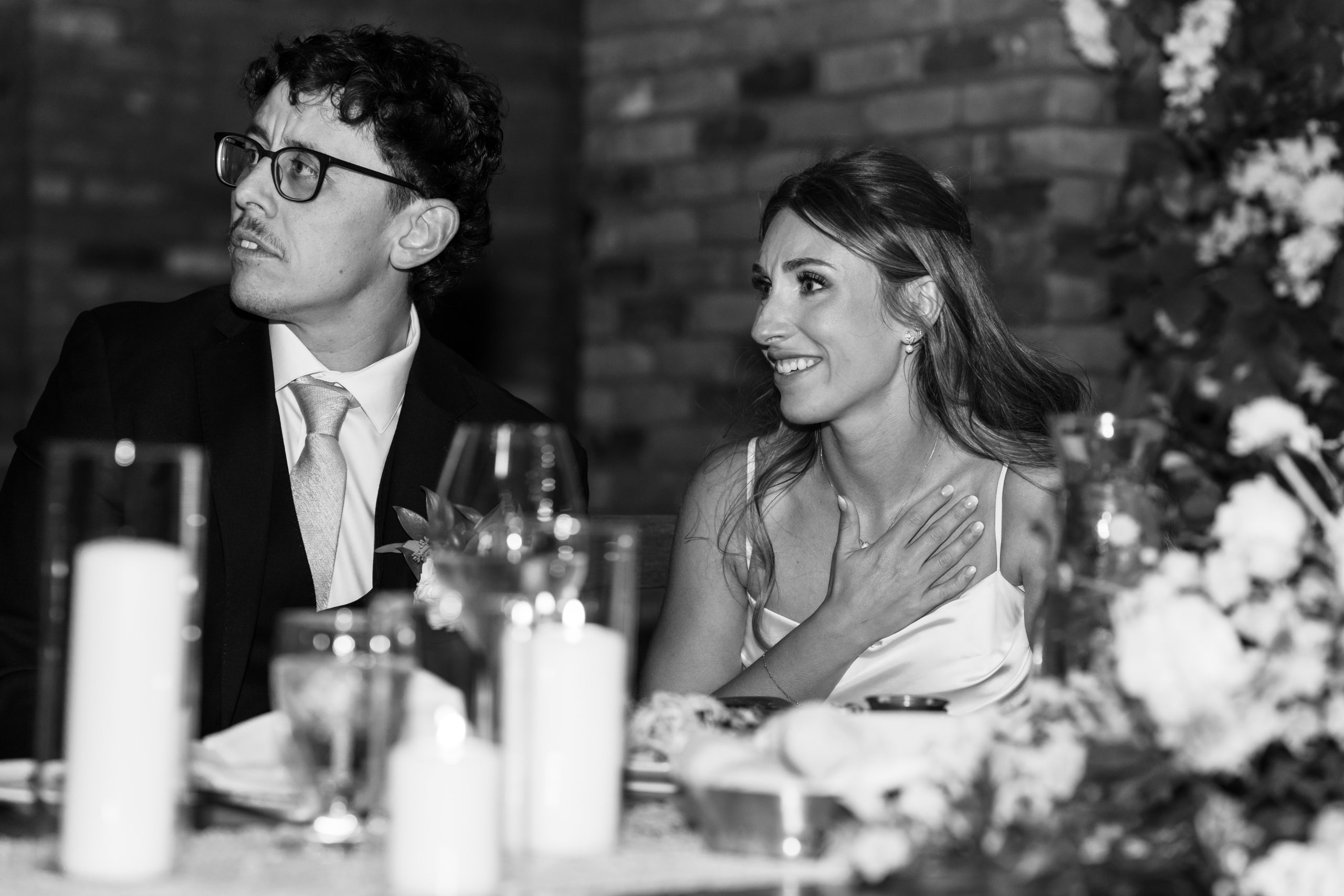 A man in a suit and tie and a woman in a white dress sit at a decorated table, smiling joyfully. Captured in Photojournalistic Wedding Photography Toronto style, the candid moment radiates celebration as she places her hand on her chest.