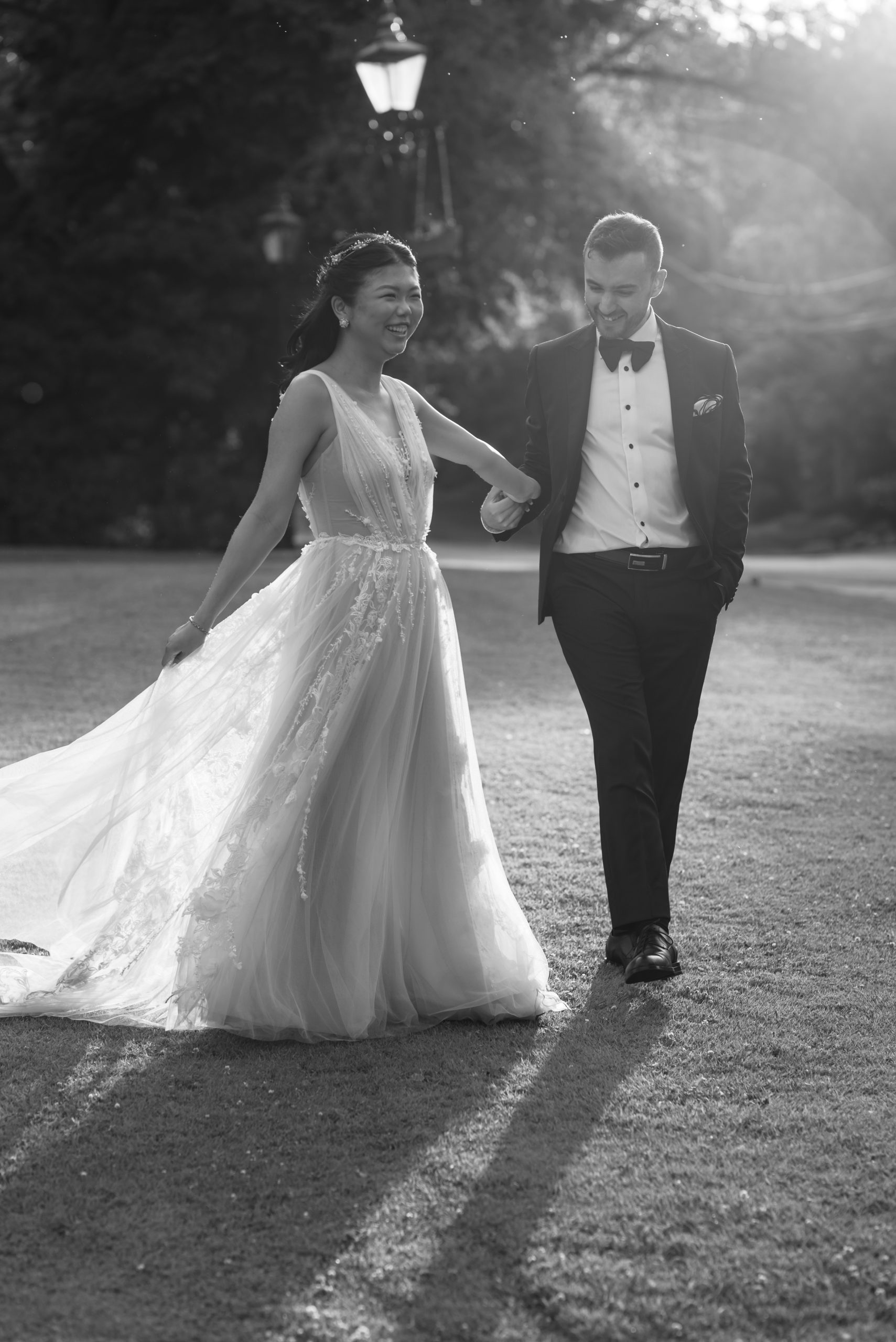 A bride and groom walk hand-in-hand outdoors, smiling at each other as their wedding photographer captures the moment. The bride’s flowing gown and the groom’s suit glow in the sunlight, creating long shadows on the grass.