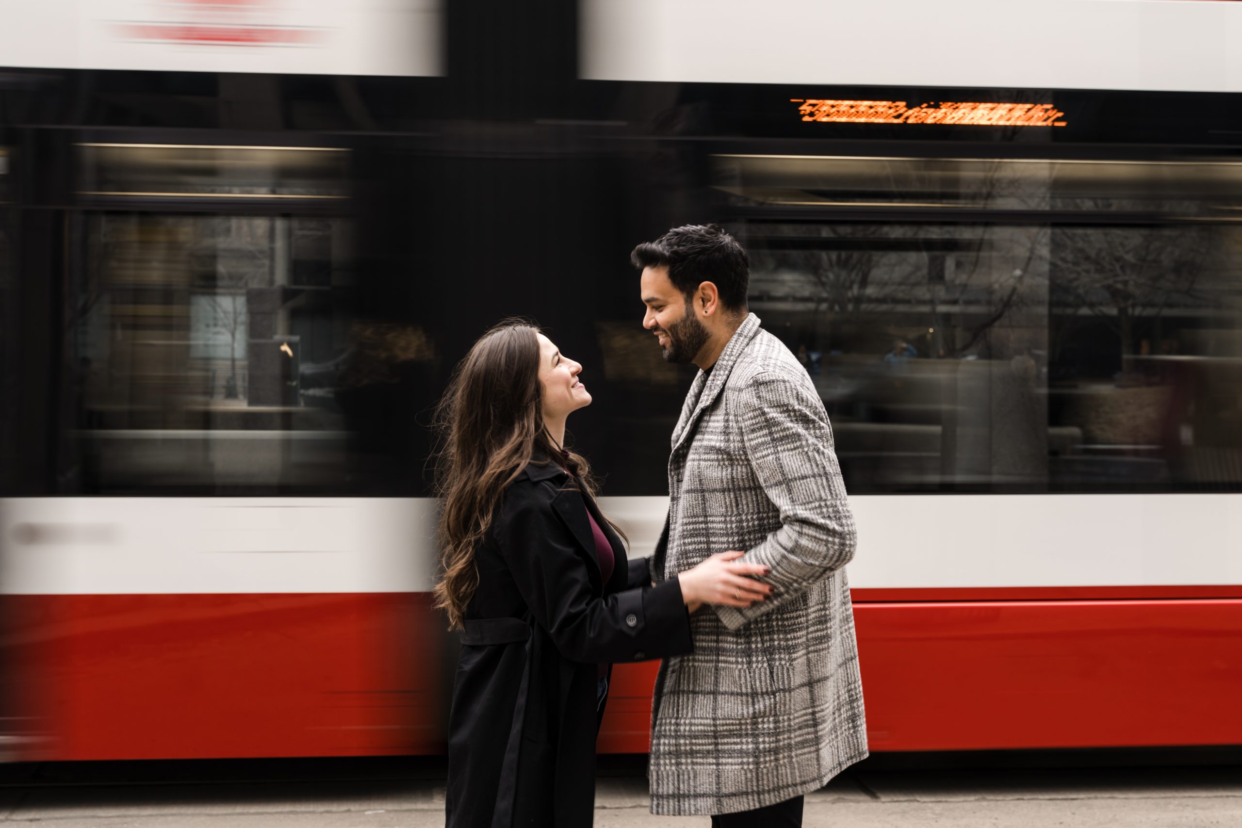 A smiling couple stands close and holds hands while looking at each other, with a blurred red and white streetcar passing by in the background. Toronto engagement photographer