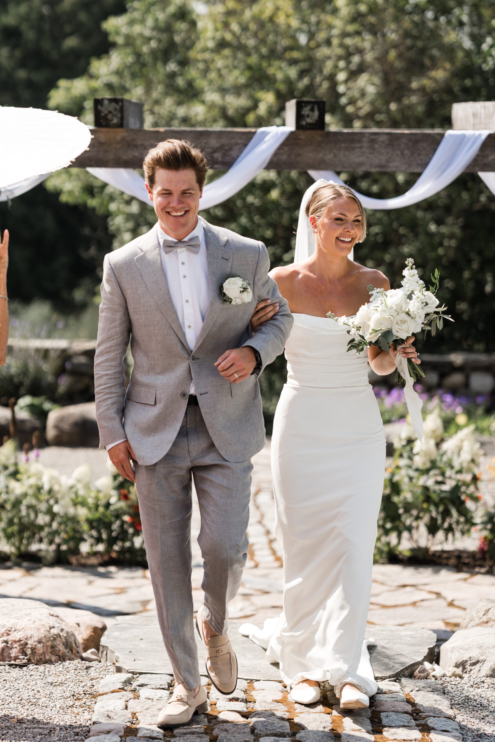 A smiling bride and groom walk arm-in-arm outdoors in an editorial style. The groom wears a light gray suit and boutonnière; the bride wears an off-the-shoulder white dress, holding a bouquet. Sunlight and greenery surround them.