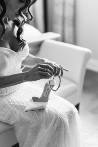 A woman in a beaded wedding dress sits on a chair, holding a high-heeled shoe and a sparkling bracelet. The image is black and white, with soft lighting highlighting her wavy hair and elegant attire.