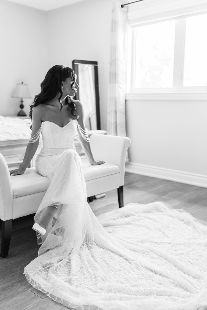 A bride in an off-the-shoulder wedding dress sits on a white bench, looking to the side. Her long train flows across the floor in a bright, softly lit room with a window and a mirror in the background.