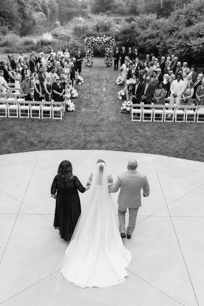 A bride walks down the aisle between two parents toward a floral arch, as wedding guests stand and watch in an outdoor ceremony, beautifully captured in an editorial wedding photography style.