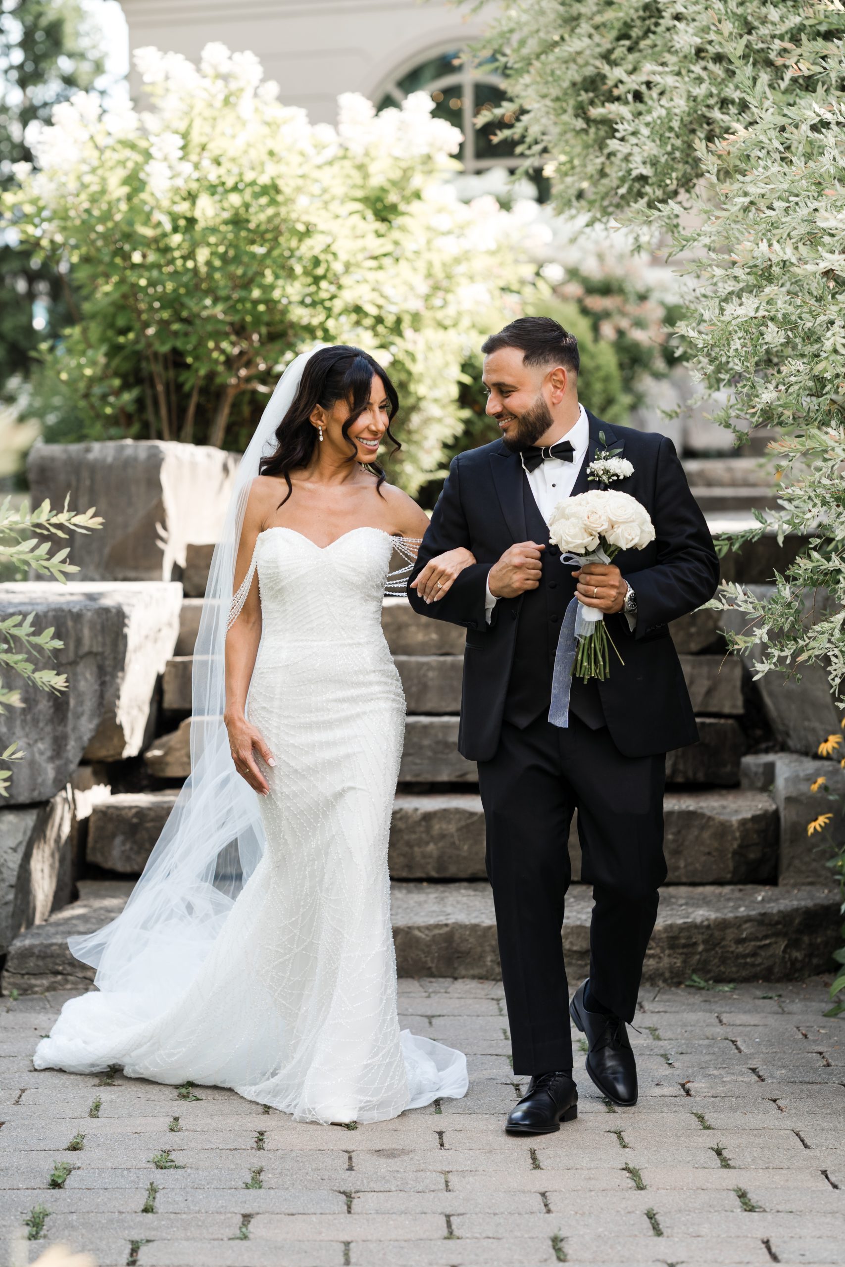 A bride in a white strapless dress and veil walks arm-in-arm with a groom in a black tuxedo holding white roses, both smiling, outdoors on a stone pathway with greenery—captured in timeless editorial wedding photography.