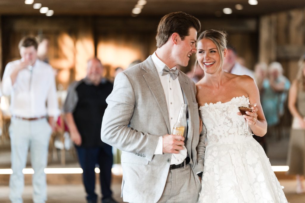 A bride in a white floral wedding dress and a groom in a light gray suit share a joyful moment while holding drinks and smiling at each other, captured in a documentary style with blurred guests in the background at a Toronto celebration.