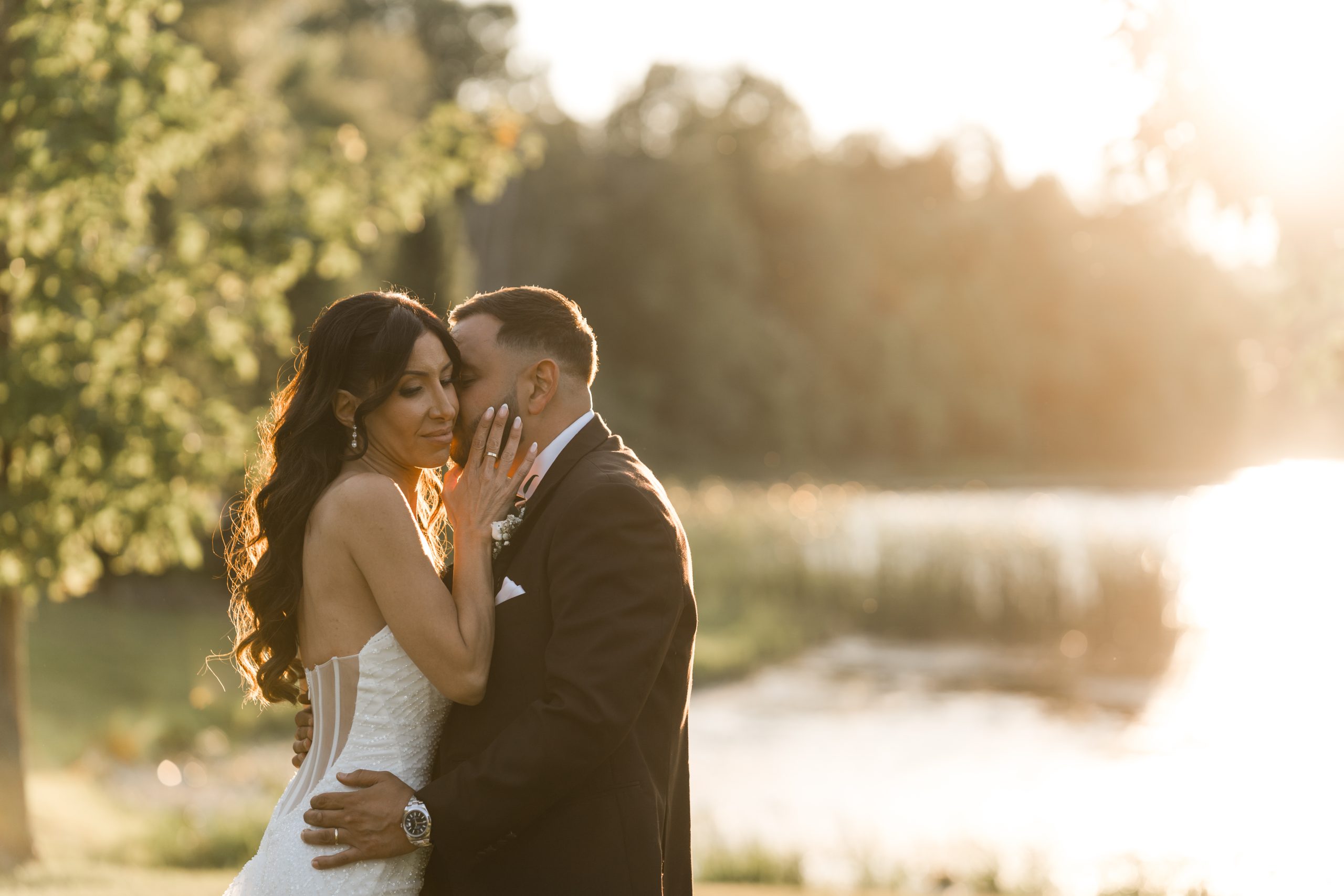 A bride and groom embrace outdoors near a sunlit lake, with trees and soft golden light in the background, creating a romantic atmosphere perfect for editorial wedding photography.