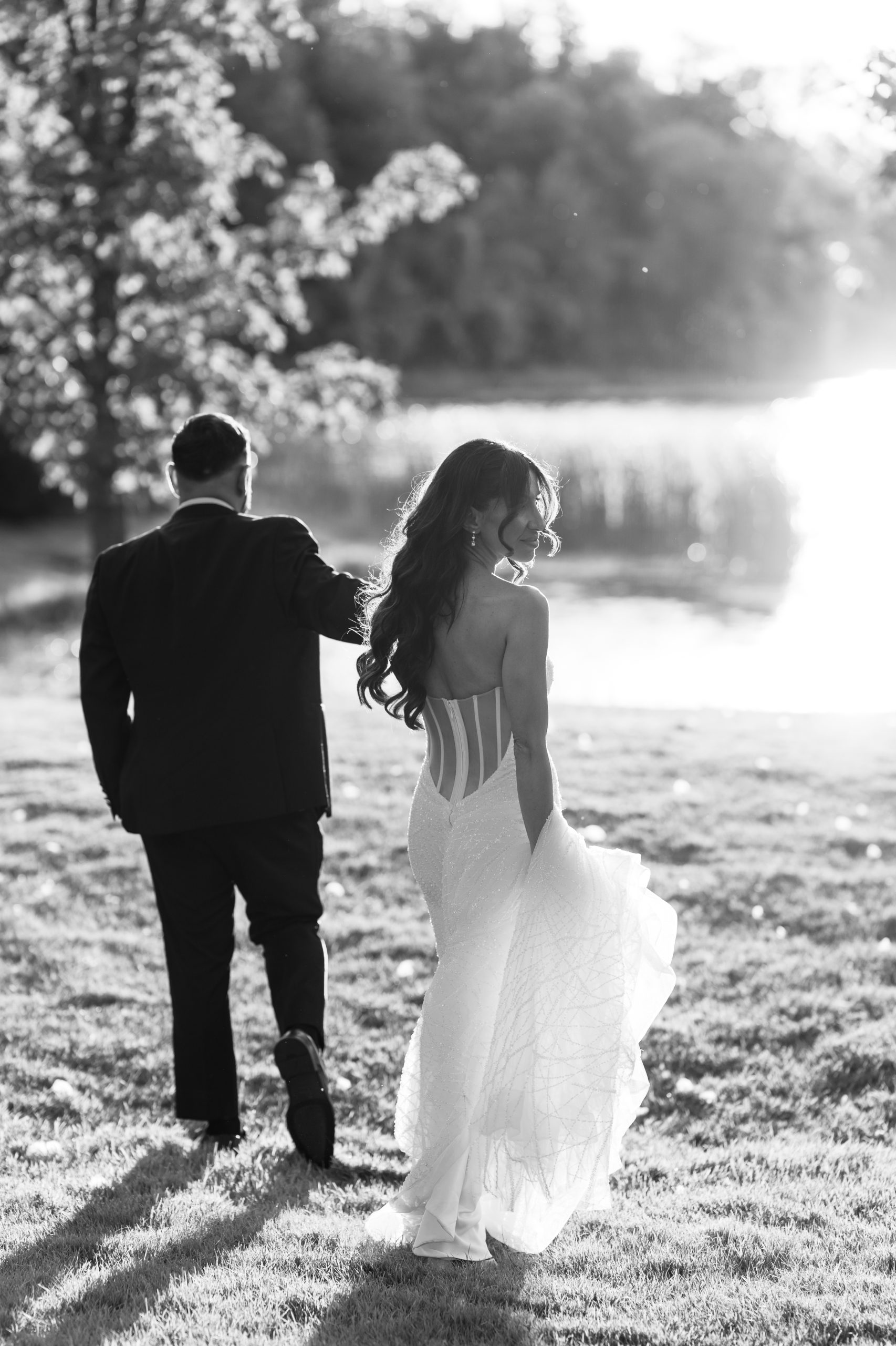 A bride in a flowing white dress and a groom in a suit walk hand in hand on grass toward a sunlit lake, surrounded by trees. The photo is black and white, with soft sunlight creating a dreamy atmosphere.