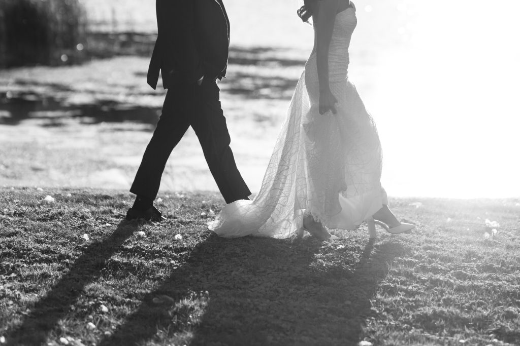 A bride and groom walk side by side outdoors, casting long shadows on the ground. The scene captures the elegance of editorial wedding photography, with sunlight creating a bright, soft glow in the background.