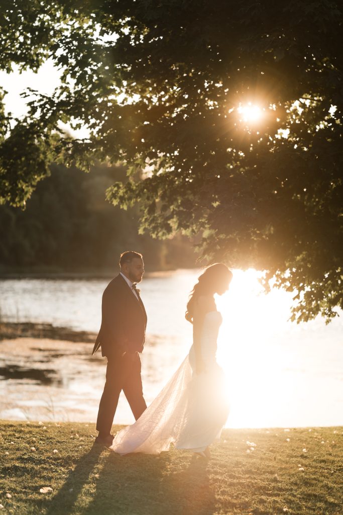 Editorial wedding photography in Toronto captures a couple dressed in wedding attire walks by a lakeside at sunset, with sunlight streaming through the trees, casting a warm glow and soft shadows on the grass.