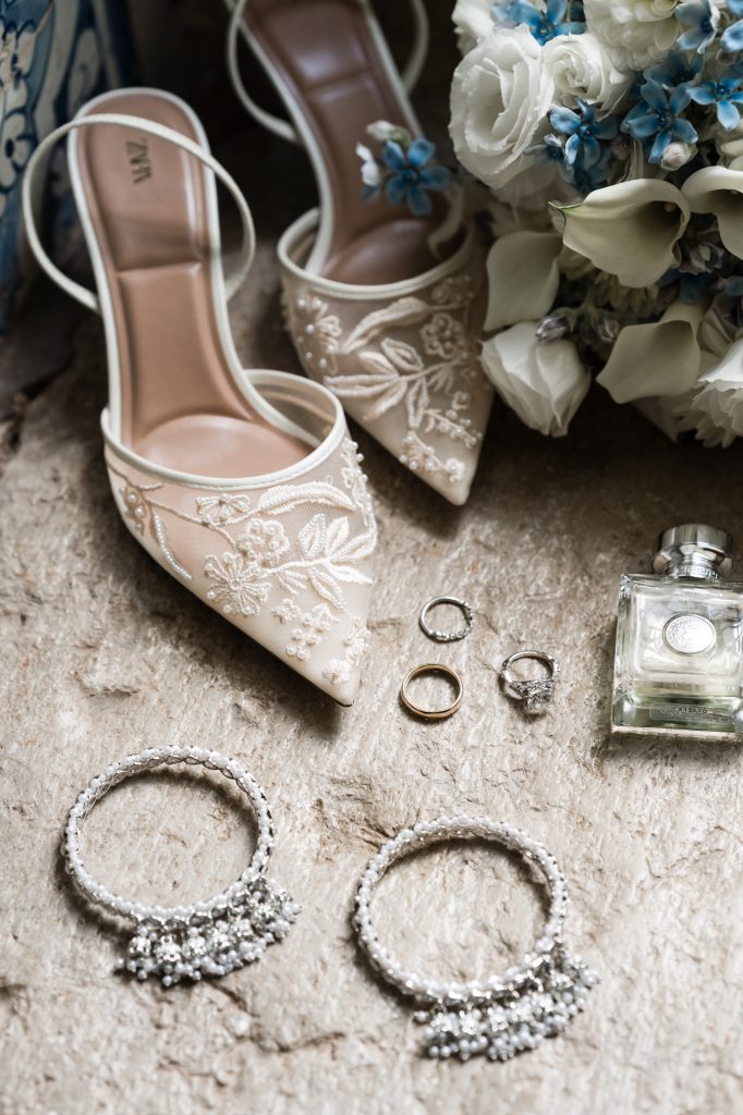 Embroidered bridal heels, a wedding bouquet, three rings, a perfume bottle, and two sparkling bracelets are arranged on a textured surface.