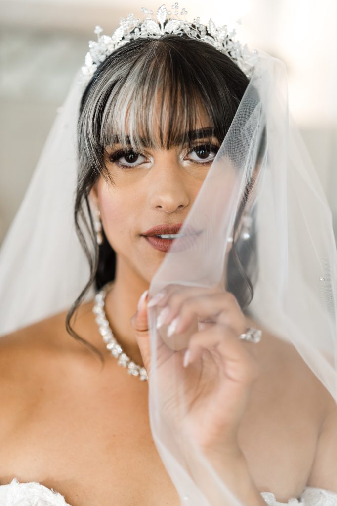 A bride with a jeweled tiara and veil holds the veil delicately, revealing her elegant makeup and jewelry, including a necklace, earrings, and a ring. She gazes softly at the camera.
