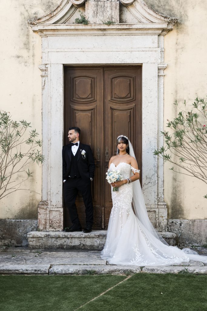 A bride in a white dress and veil holds a bouquet, standing next to a groom in a black suit leaning against a large wooden door set in a stone building, with plants on either side.