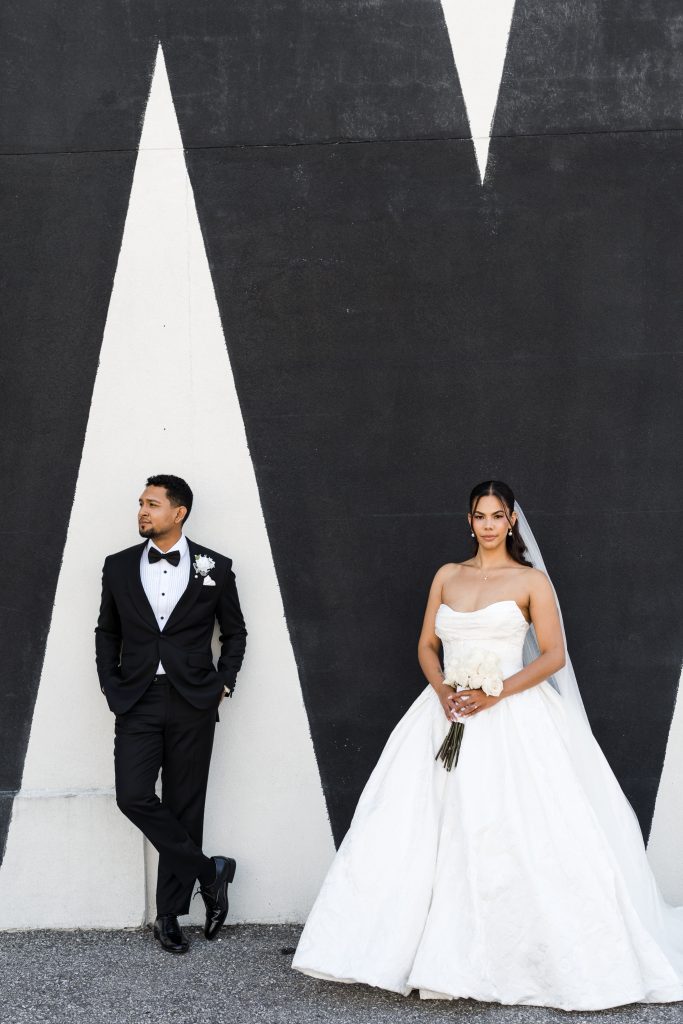 A bride in a strapless white gown and veil holds flowers, standing beside a groom in a black tuxedo. They pose against a wall with bold black and white geometric patterns.