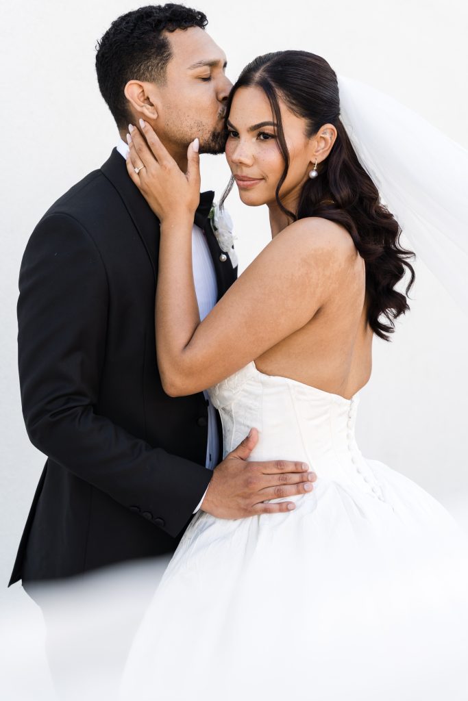 A groom in a black suit gently kisses the forehead of a bride in a strapless white wedding dress and veil, while holding her waist. The bride smiles softly, holding his face, against a plain white background.