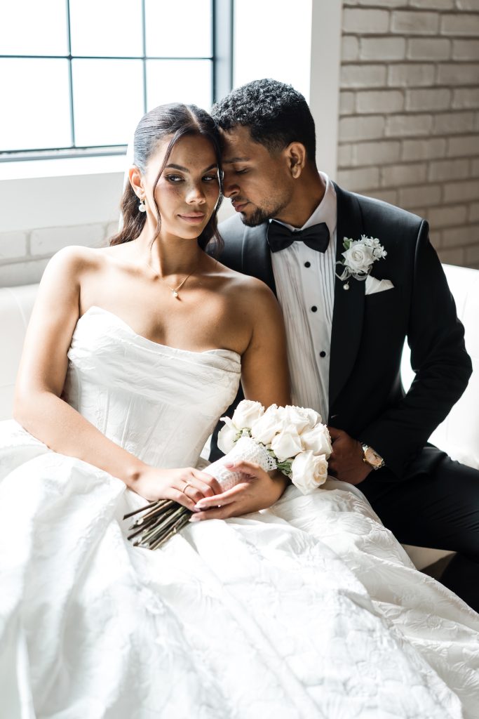 A bride in a white strapless gown holds a bouquet of white roses while sitting beside a groom in a black tuxedo. They sit close together on a white couch, sharing an intimate, affectionate moment.