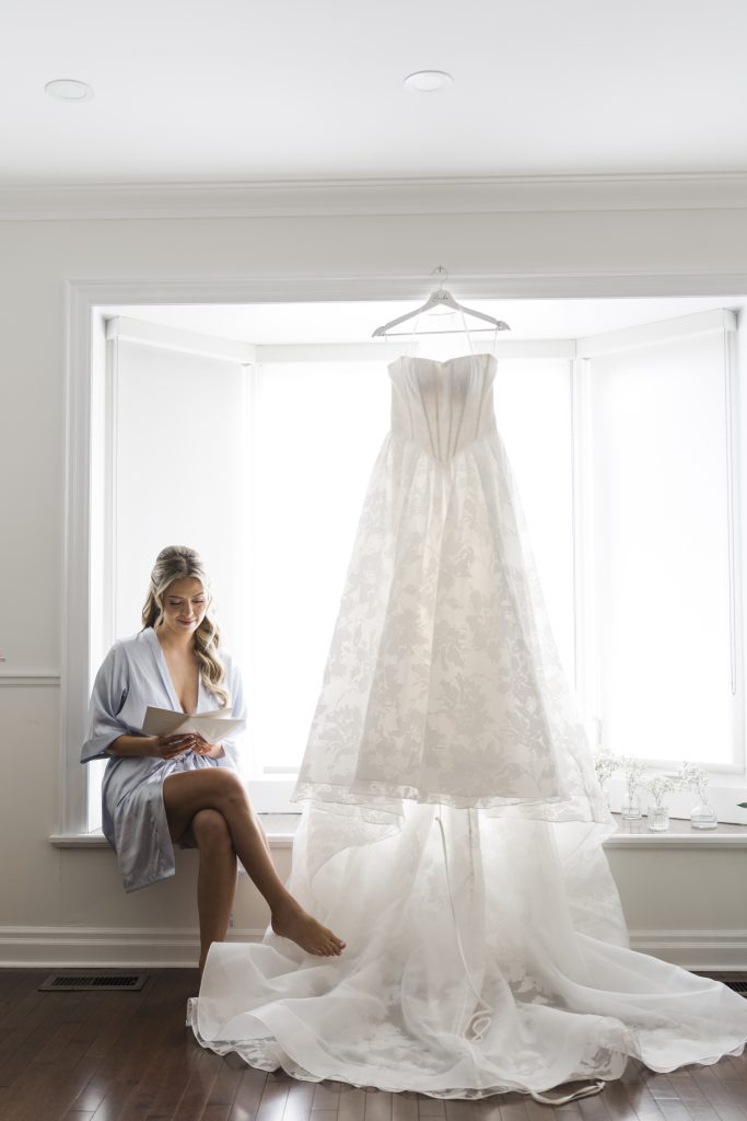 A woman in a light robe sits by a large window, reading a card. A white wedding dress hangs on a hanger in the bright window next to her, softly lit by natural light. Captured by a Toronto wedding photographer