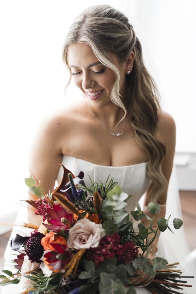 A bride with long, wavy blonde hair and a strapless white dress smiles down at a vibrant bouquet of flowers, featuring red, pink, and purple blooms with assorted greenery, against a bright, softly-lit background. Image taken by a Toronto wedding photographer