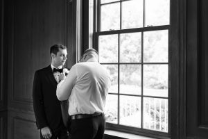 A man in a tuxedo stands by a large window while another adjusts his boutonnière. Natural light fills the room, giving the scene an editorial quality perfect for photography, and creating a calm and focused atmosphere.