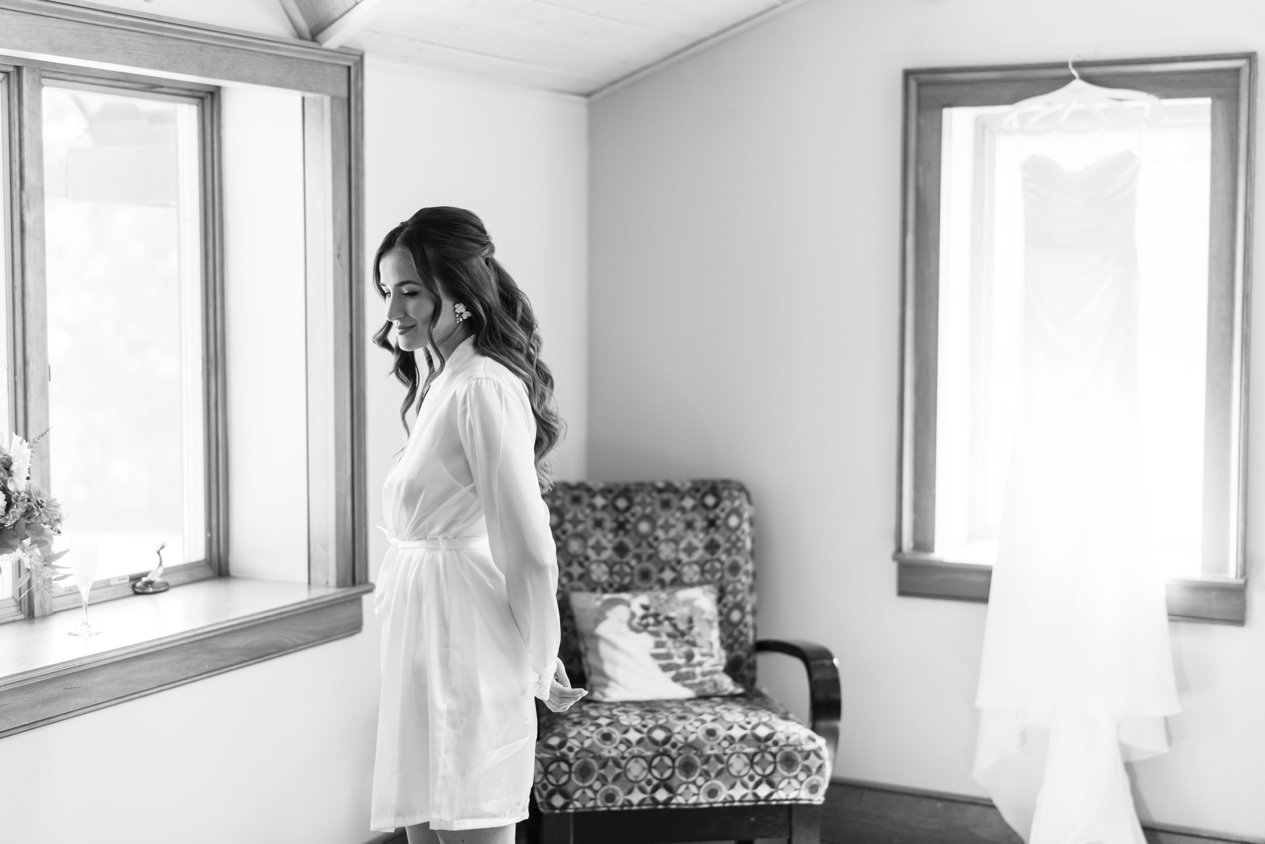 A woman in a robe stands by a bright window, smiling and looking down. In the softly lit, editorial-style black and white image, a wedding dress hangs near a patterned chair in the background of this Toronto room.