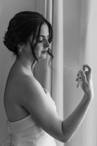 A woman in a strapless dress holds and sprays a perfume bottle near her neck, standing by a curtain. This black and white editorial image captures an intimate, documentary-style moment reminiscent of classic Toronto photography.