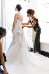 A bride in a strapless white gown stands as two women in green dresses help adjust her train and tie the large bow at the back—capturing a softly lit, editorial moment in photography.