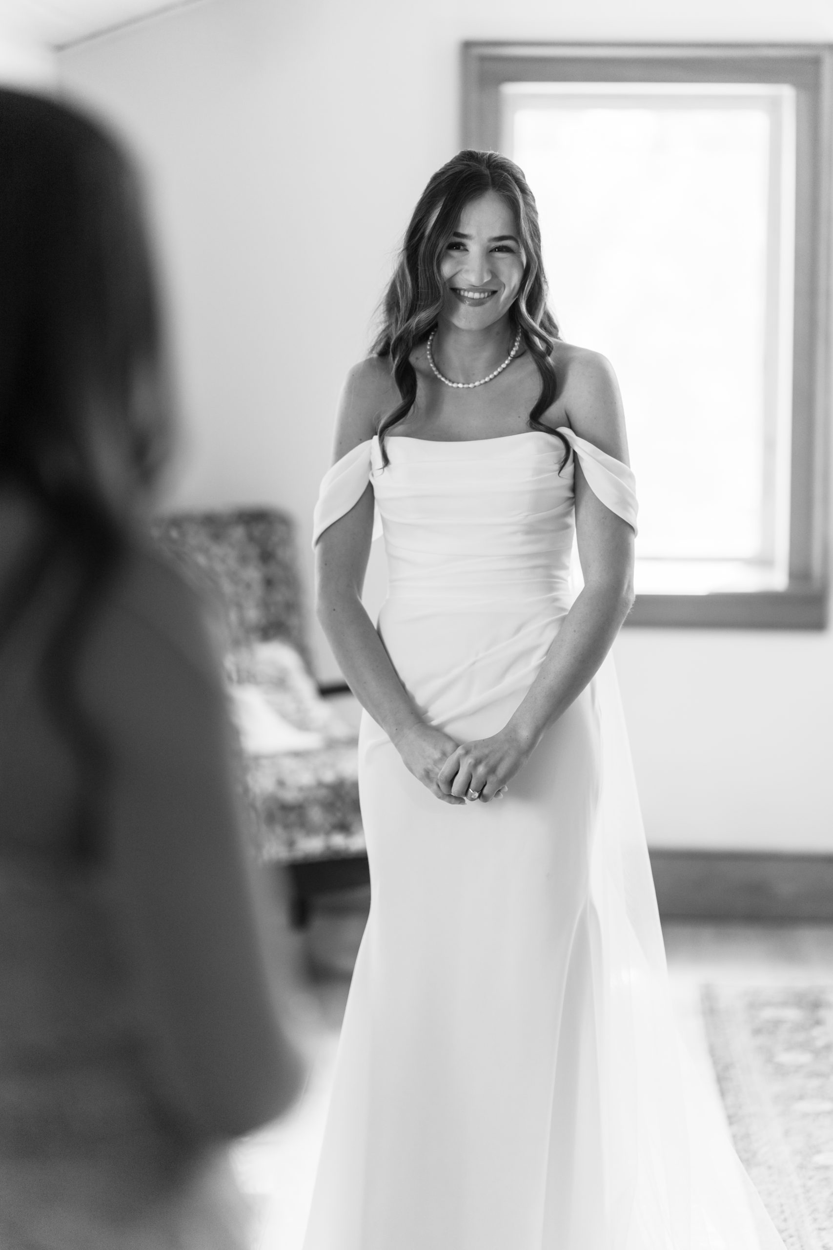 A smiling bride in an off-the-shoulder wedding dress stands indoors near a window, looking at someone out of focus in the foreground. This black and white editorial photograph captures a timeless, intimate moment.
