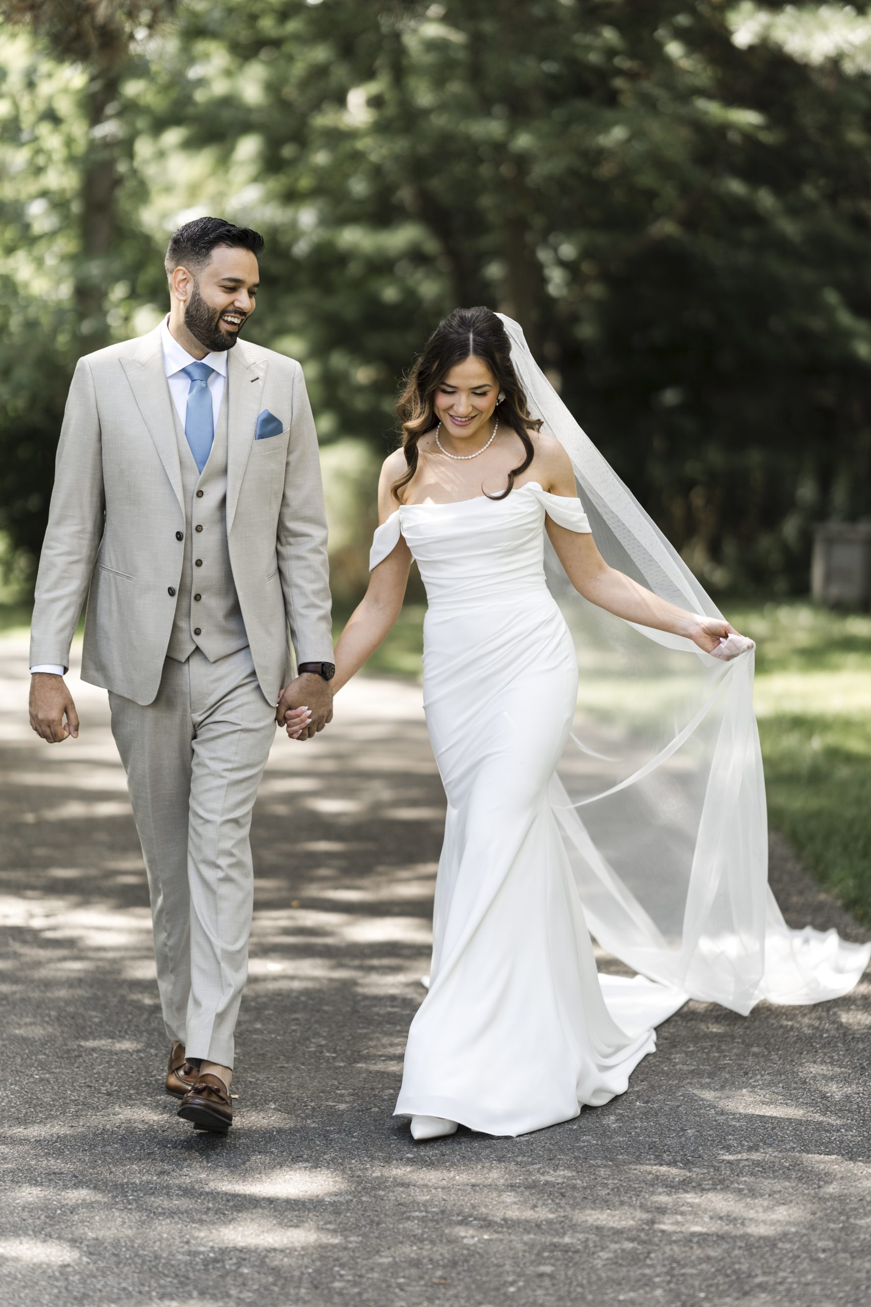 A bride in a white off-the-shoulder gown and veil walks hand in hand with a groom in a light beige suit and blue tie, smiling together on a sunlit path, creating a perfect moment for stunning editorial photography surrounded by greenery.