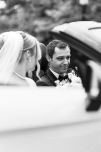 A bride and groom sit together in a convertible car. The bride wears a veil and pearl necklace, and the groom smiles gently in his tuxedo. Captured in black and white, this editorial image by a Toronto photographer features a beautifully blurred background.