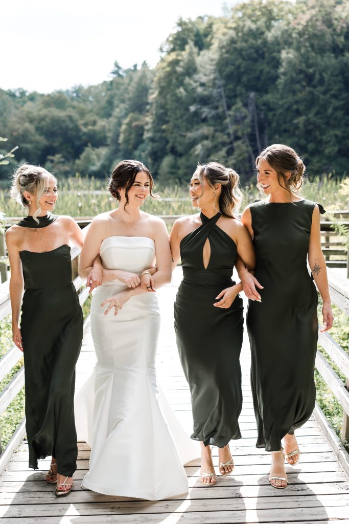 A bride in a white strapless gown walks on a wooden path outdoors, smiling and linking arms with three bridesmaids in matching dark green dresses. Lush greenery and trees fill the background, creating an editorial-worthy Toronto wedding scene.