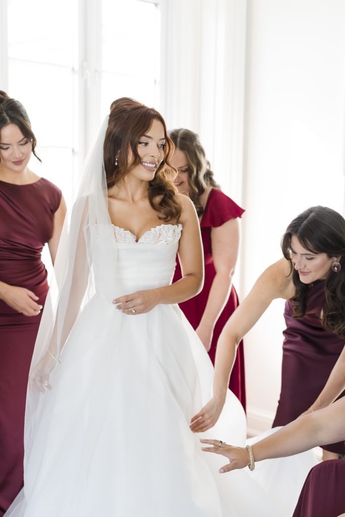 A smiling bride in a white wedding dress and veil is surrounded by four bridesmaids in matching burgundy dresses, helping her with her gown in a bright Toronto room—perfect for timeless documentary photography.