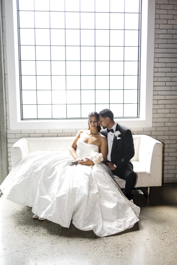A bride in a white gown and a groom in a black tuxedo sit closely on a white couch, smiling and leaning into each other, with a large window behind them letting in natural light. Captured by a Toronto wedding photographer