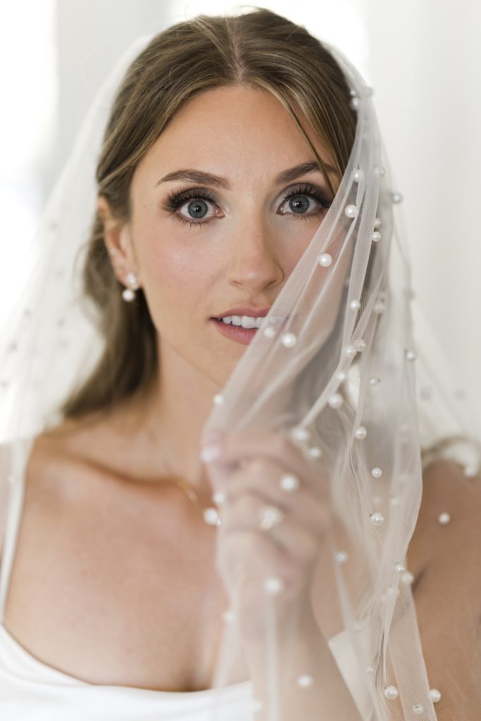 A bride with long brown hair holds a sheer, pearl-dotted veil over part of her face. Captured by an editorial wedding photographer Toronto, she has natural makeup, wears pearl earrings, and gazes softly at the camera.