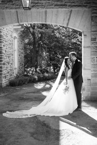 A bride and groom stand under a stone archway, smiling and embracing on their wedding day. Sunlight streams through as a Toronto photographer captures this romantic, black-and-white scene with the bride in a long veil holding her bouquet.