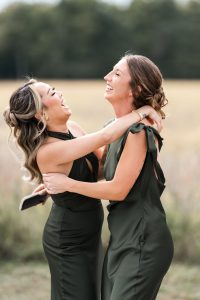 Two women in matching olive green dresses laugh and embrace outdoors, standing in a field with blurred trees behind them. One holds a phone as they share a joyful moment captured in a candid, editorial wedding style.