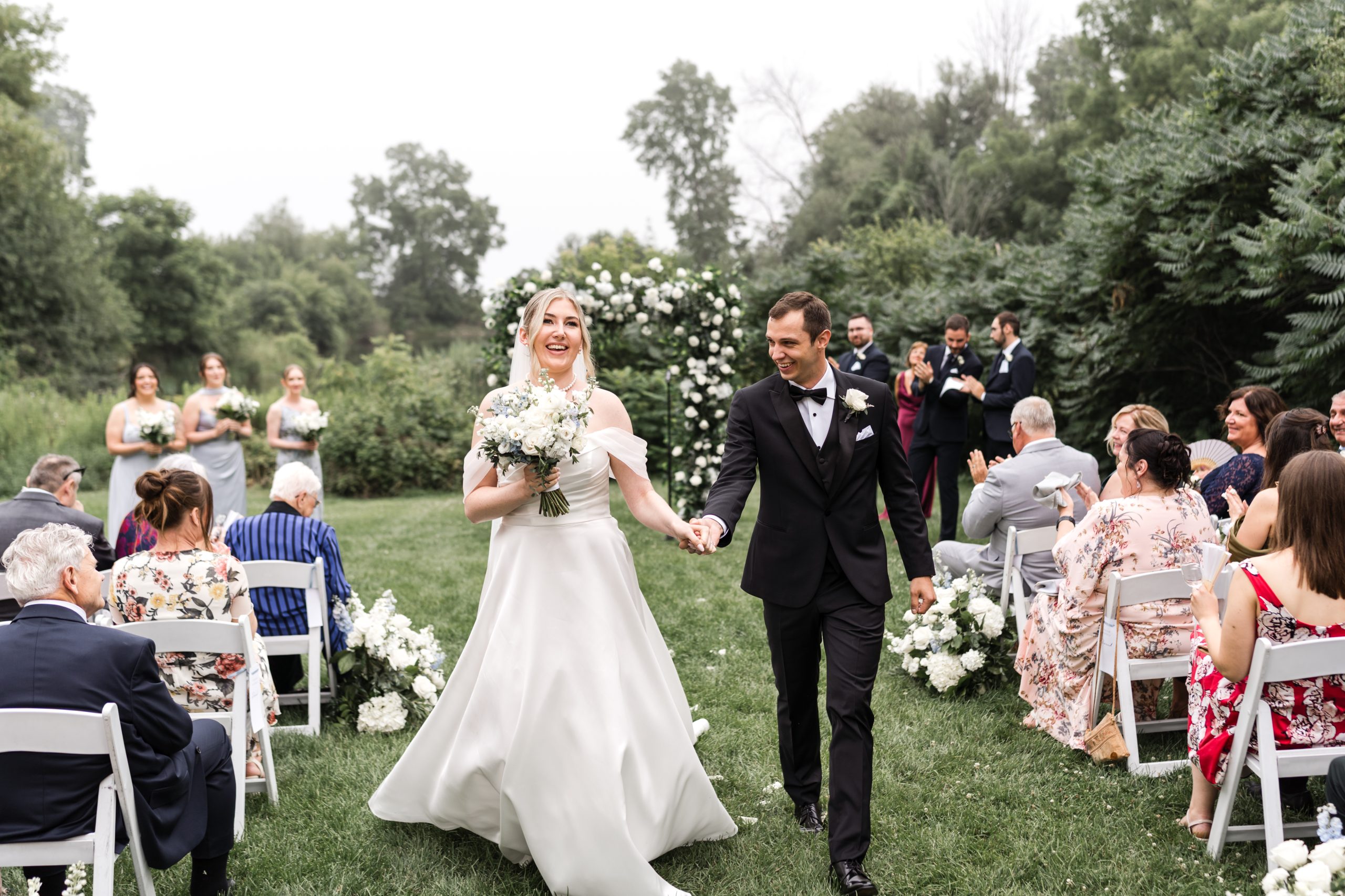 A bride and groom walk hand in hand down the aisle at their Toronto wedding, smiling joyfully. Guests clap and celebrate while bridesmaids stand near a floral arch, surrounded by greenery in this documentary-style moment.