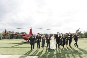 A bride and groom walk with their wedding party on a grassy field, all dressed formally in black and white, with a red helicopter parked in the background under a cloudy sky—captured in a documentary photography style near Toronto.