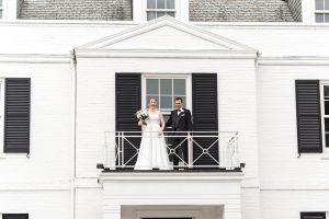 A bride and groom stand side by side on a small balcony of a white brick building with black shutters, smiling and facing forward. Captured in elegant editorial photography, the bride holds a bouquet in her white dress; the groom is in a black tuxedo.
