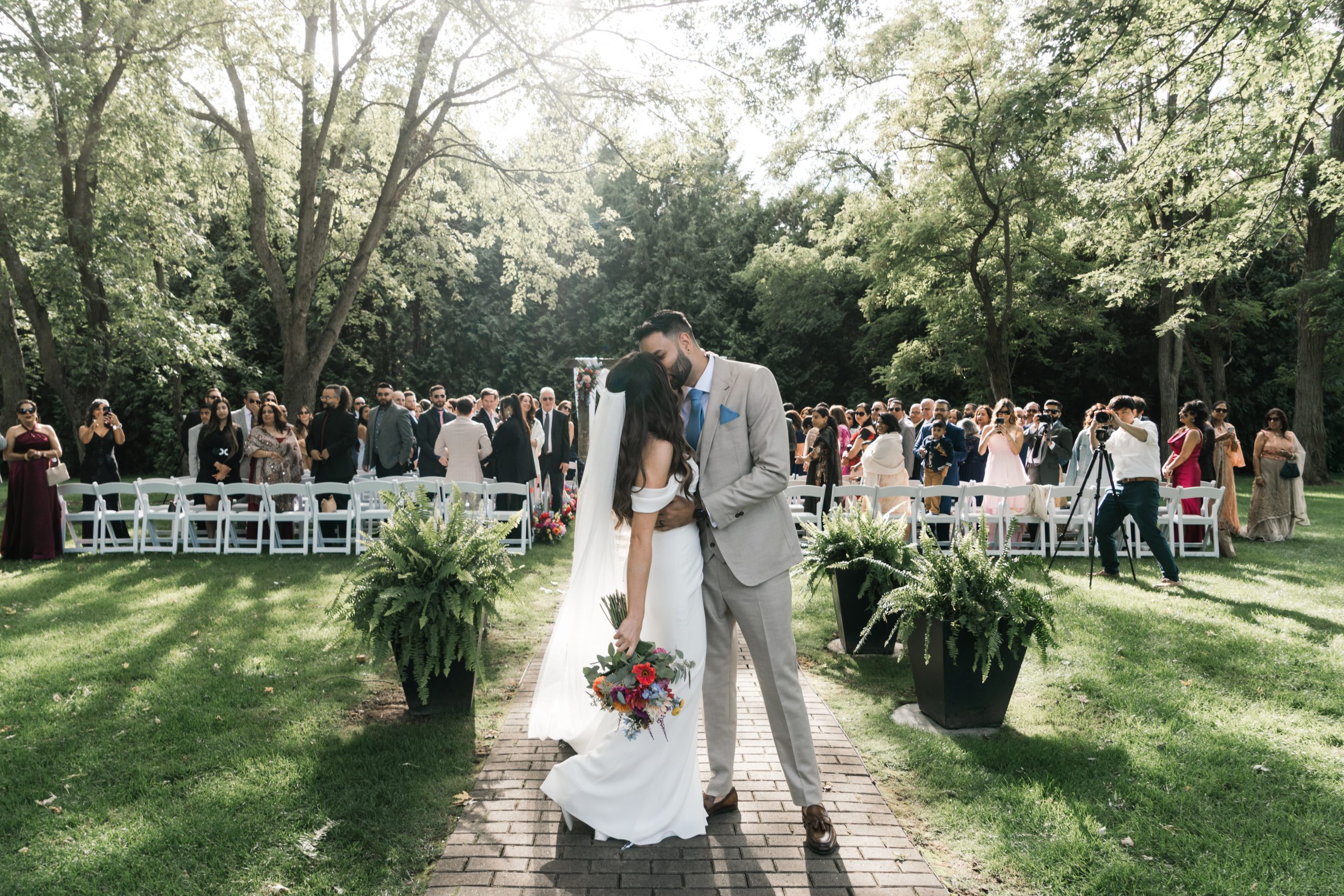 A bride and groom share a kiss outdoors on a sunlit walkway in Toronto, surrounded by lush greenery and wedding guests seated in white chairs. The editorial-style photo captures her in a white dress with a bouquet and him in a light suit.