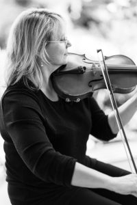 A woman with shoulder-length blonde hair and glasses plays a violin, holding the instrument under her chin and drawing a bow across the strings. Captured in black and white, the image has a documentary feel, as if seen through a photographer’s lens.